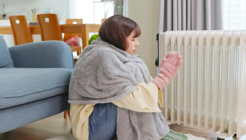 Ragazza avvolta in una coperta, con guanti rosa, vicino a un termosifone acceso, ma la casa è fredda nonostante i termosifoni.