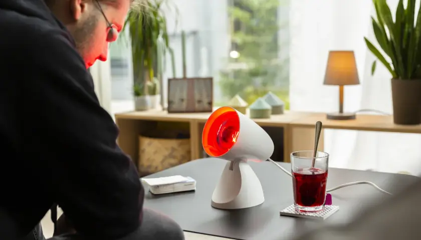 Mann mit rotlicht bei fieber, der sich auf eine Wärmelampe konzentriert. Daneben ein Glas mit rotem Getränk.