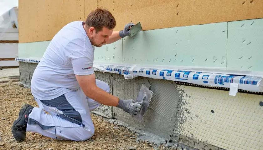 Handwerker kniet und verputzt die Sockelzone einer Hausfassade mit einer Kelle.