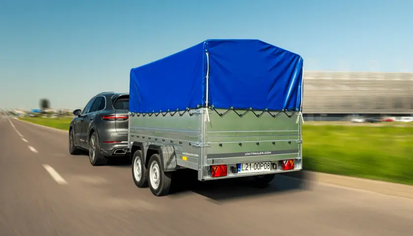 Przyczepa kempingowa bez prawa rejestracji, z niebieską plandeką, ciągnięta przez ciemny SUV.