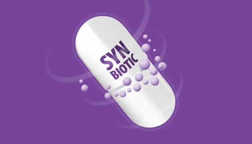 Biała kapsułka z napisem "SYN BIOTIC" na fioletowym tle. Drobne kuleczki unoszą się wokół. Dowiedz się, synbiotyk co to.