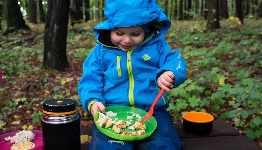 Małe dziecko w niebieskiej kurtce cieszy się posiłkiem w lesie. To idealne jedzenie w góry, które dodaje energii do dalszych przygód.