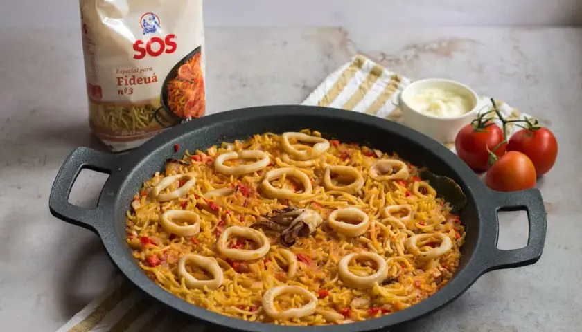 Fideuá con calamares lista para servir, acompañada de tomates y alioli.