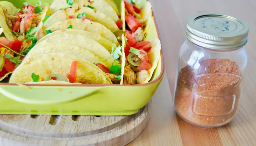 Domowa przyprawa do taco w słoiku obok talerza pełnego tacos z pomidorami i jalapeno. Lepsza niż gotowa z torebki!