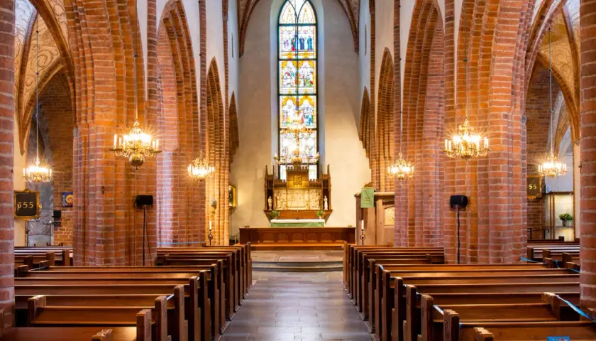 Interiör av Helga Trefaldighets kyrka med bänkar, tegelvalv och ett färgstarkt glasmålat fönster bakom altaret.