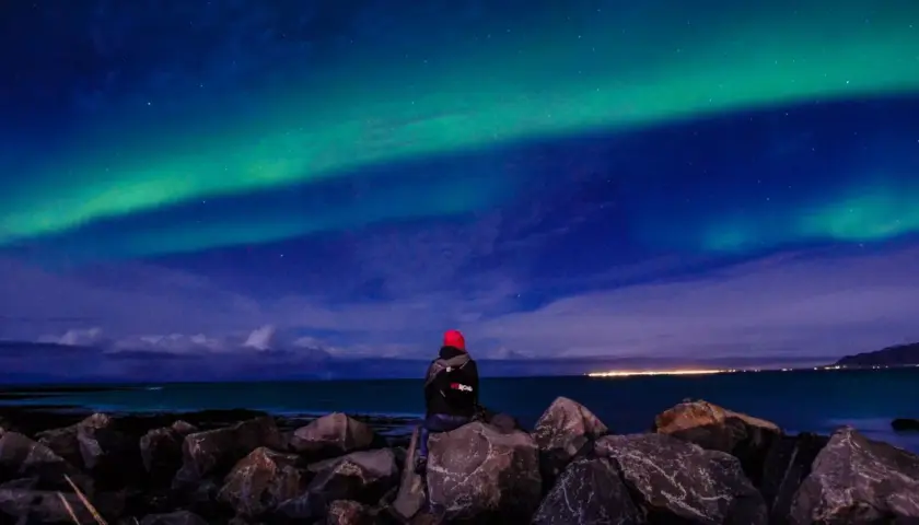 Person sitzt auf Felsen am Meer und blickt auf Nordlichter. Perfektes Wetter auf Island im September für dieses Naturschauspiel.