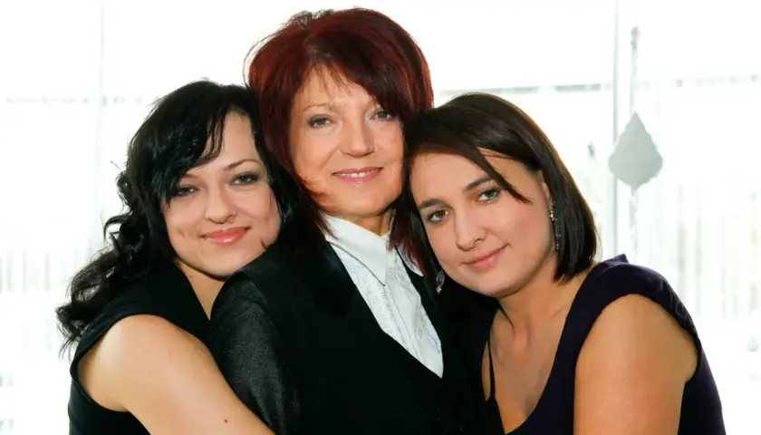 Trzy kobiety: starsza z rudymi włosami w czarnej marynarce i dwie młodsze, córki, obejmujące ją.