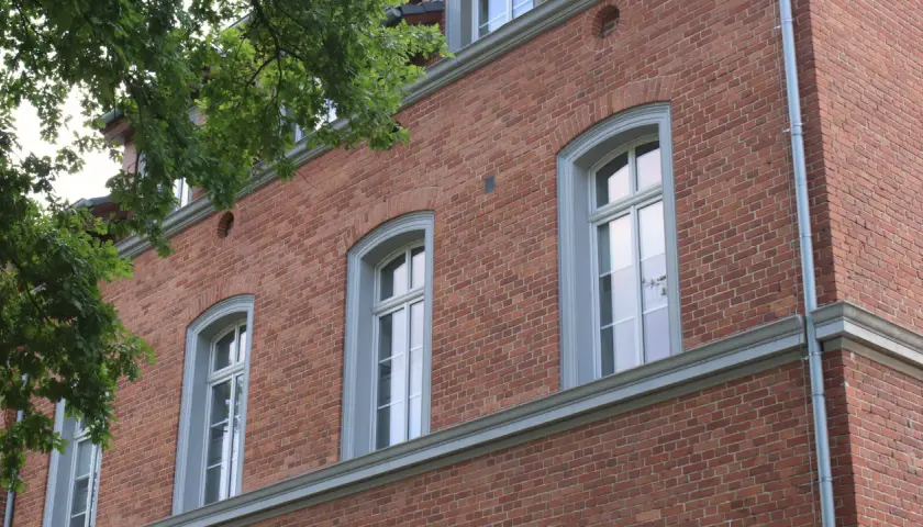 Eleganckie okna drewniane zabytkowe w ceglanej kamienicy, otoczone zielenią drzew.