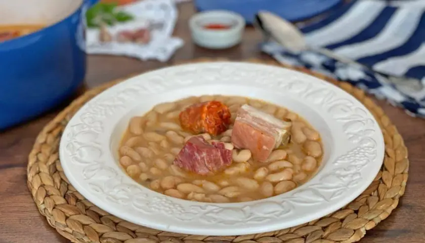 Plato de judías blancas con chorizo y tocino, servido en una vajilla blanca sobre una esterilla de mimbre.