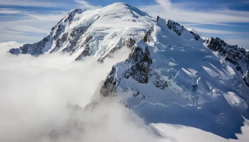 Le Mont Blanc, majestueux et enneigé, émerge des nuages. Des alpinistes escaladent ses pentes glacées sous un ciel bleu éclatant.