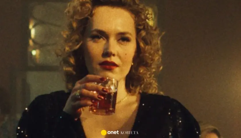 Kobieta z burzą blond loków i czerwoną szminką, pijąca drinka. W tle widać fragmenty scenografii, sugerujące klimat filmu "Jak pokochałam gangstera obsada".