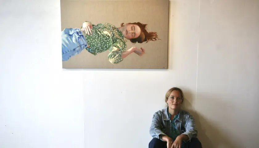 Konstnären Elin Boberg sitter på golvet framför sin målning av en sovande kvinna i grönt mönstrat linne och blå jeans.