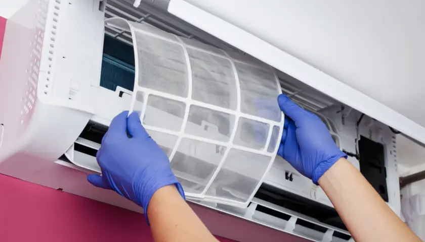 Manos con guantes azules retiran el filtro del aire acondicionado. ¿Para qué sirve? Para atrapar polvo y alérgenos, manteniendo el aire limpio.