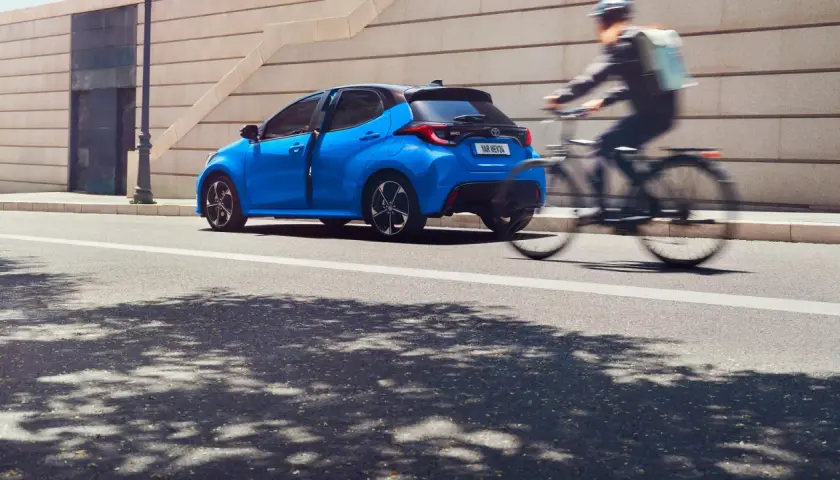 Una Toyota Yaris blu brillante sfreccia in città, mentre un ciclista con zaino passa velocemente. Diversi modelli di Toyota Yaris sono disponibili per ogni stile di vita.