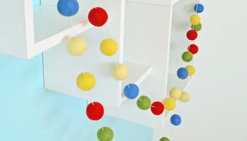 Eine bunte Filzgirlande mit Kugeln in Rot, Blau, Gelb und Grün hängt an weißen Regalen.