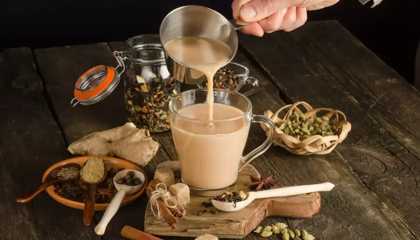 Ręka wlewa mleko do kubka, tworząc aromatyczną herbatę po irlandzku. Wokół przyprawy: cynamon, kardamon, imbir.