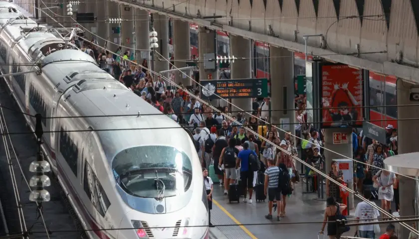 Un tren AVE espera en la estación, listo para llevar pasajeros de Lleida a Sevilla. Gente con maletas se agolpa en el andén.
