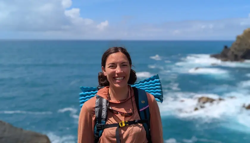 A smiling hiker with a backpack and sleeping pad enjoys the view of the ocean waves crashing on the shore along the Fischerweg trail in Portugal.