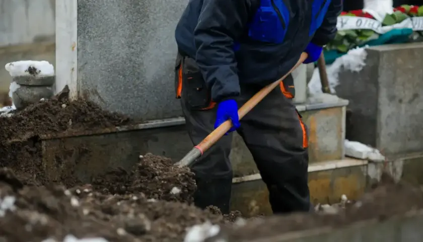 Grabarz w niebiesko-czarnym ubraniu kopie grób łopatą. W tle widać nagrobki i wieńce pogrzebowe.