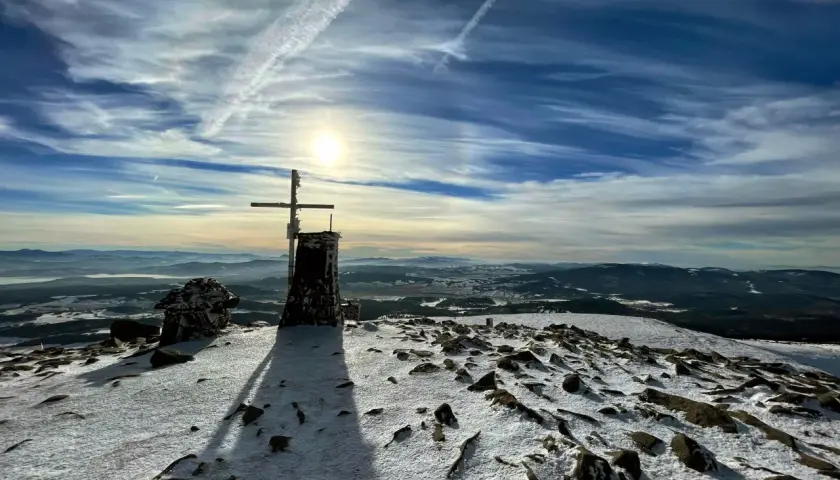 Szczyt Babiej Góry zimą. Krzyż i kamienna konstrukcja na ośnieżonym szczycie. W tle rozległa panorama gór i niebo z chmurami. Szlaki Babiej Góry w śniegu.