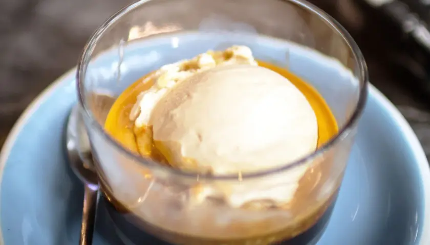Pyszne affogato: gałka lodów waniliowych w gorącym espresso, podana w szklance na jasnoniebieskim talerzyku.