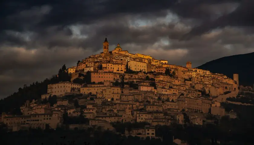 Antico borgo di Trevi, Umbria, illuminato da un sole dorato che filtra tra nuvole scure. Case in pietra si aggrappano alla collina.