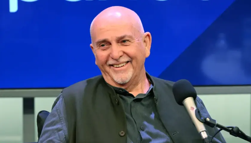 Uśmiechnięty Peter Gabriel, legendarny wokalista, w studiu. W tle niebieskie logo, przypominające o jego bogatej karierze z zespołem Genesis.