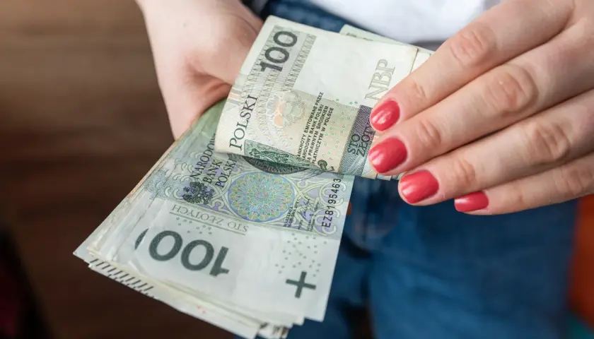 Dłonie trzymające stos banknotów 100 zł. Porównanie zasiłku stałego a renty socjalnej.