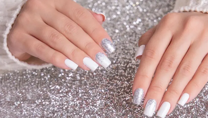 Dłonie z białym french manicure i brokatem na paznokciach, na tle srebrnego brokatu.
