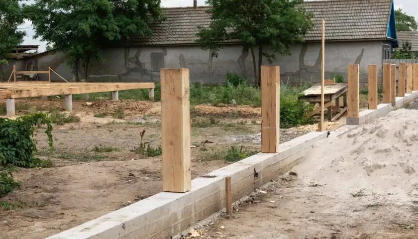 Solidny fundament pod ogrodzenie z drewnianymi słupkami w trakcie budowy. W tle widać dom i konstrukcję drewnianą.