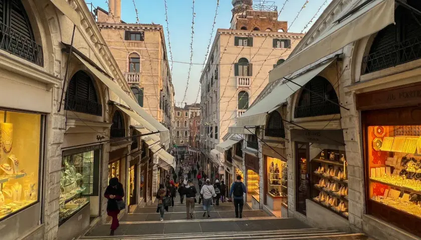 Schody na moście Rialto w Wenecji, z witrynami sklepów z biżuterią po bokach. Tłum ludzi schodzi w dół.