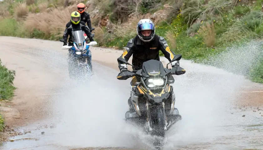 Motociclistas en un **conti trail attack 4 test** atraviesan un charco de agua en un camino de tierra, levantando una gran estela.