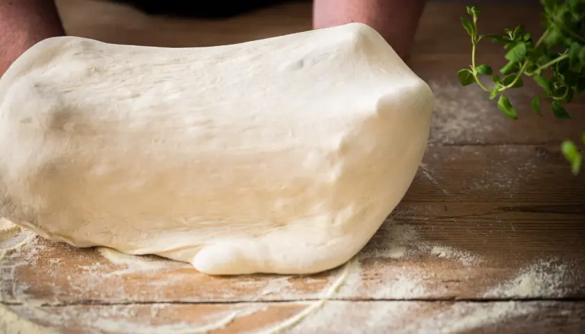 Frischer Teig für Pizza Semola wird auf einem Holztisch geknetet, bestäubt mit Mehl. Kräuter liegen daneben.