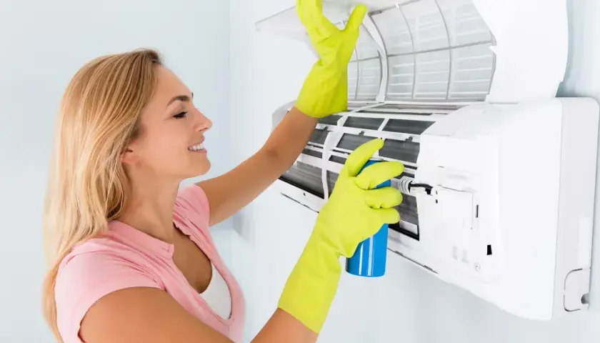Mujer con guantes verdes limpia el aire acondicionado que huele a vinagre, rociando un producto para eliminar olores.