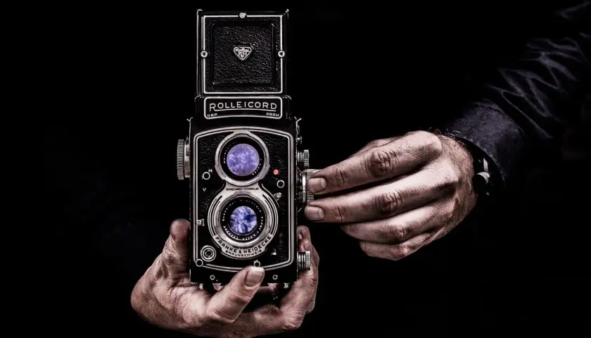 Dwie dłonie trzymają zabytkowy aparat Rolleiflex, którego obiektywy odbijają światło, tworząc wrażenie złotych proporcji w kadrze.