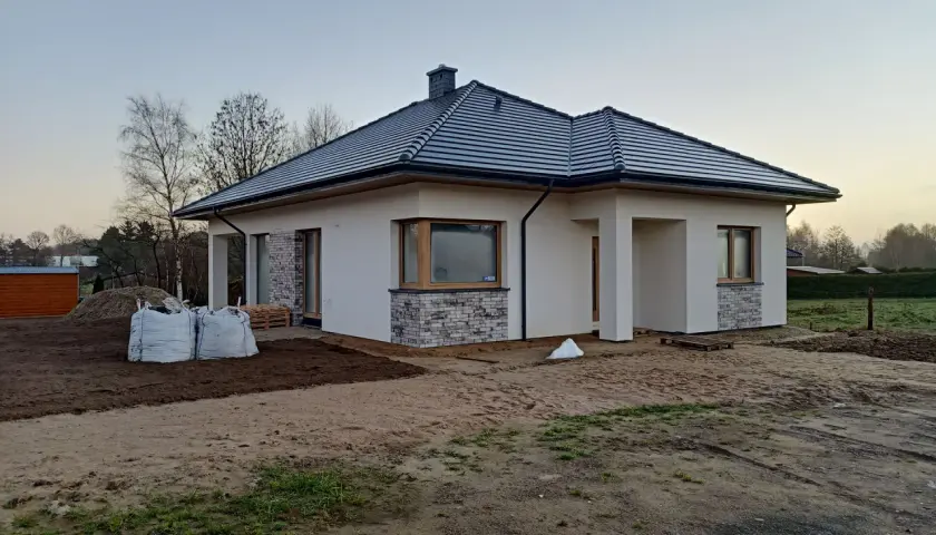 Nowy, nowoczesny dom w renklodach 15 z szarym dachem i białymi ścianami, otoczony placem budowy.