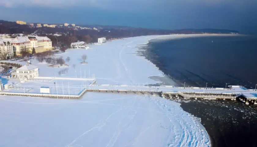 Zimowy spacer po molo, gdzie zimą nad morze przyciąga biel śniegu i lodowe krajobrazy. Widok na zamarznięte plaże i budynki w oddali.