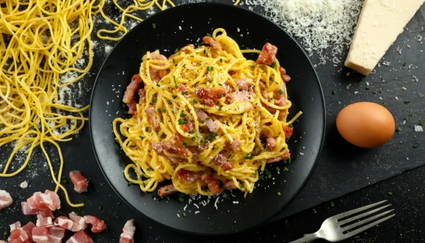 Pyszne spaghetti carbonara na talerzu, obok surowy boczek, jajko i ser. Zastanawiasz się, ile kalorii ma spaghetti carbonara?