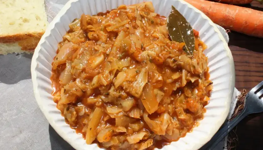 Pyszny bigos z młodej kapusty w białej misce, obok kromka chleba i marchewki.