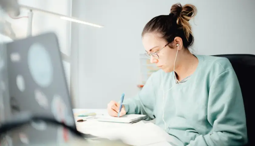 Student med hörlurar skriver i anteckningsbok vid skrivbordet, inspirerad av sitt universitetsutbildning i kreativt skrivande.