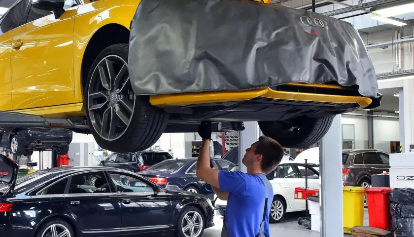 Mechanik pracuje pod żółtym Audi, sprawdzając, ile kosztuje wymiana oleju silnikowego w samochodzie. W tle inne auta.