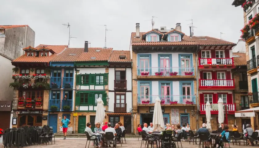 Fachada de casas coloridas en Hondarribi, con balcones floridos y gente disfrutando en terrazas.