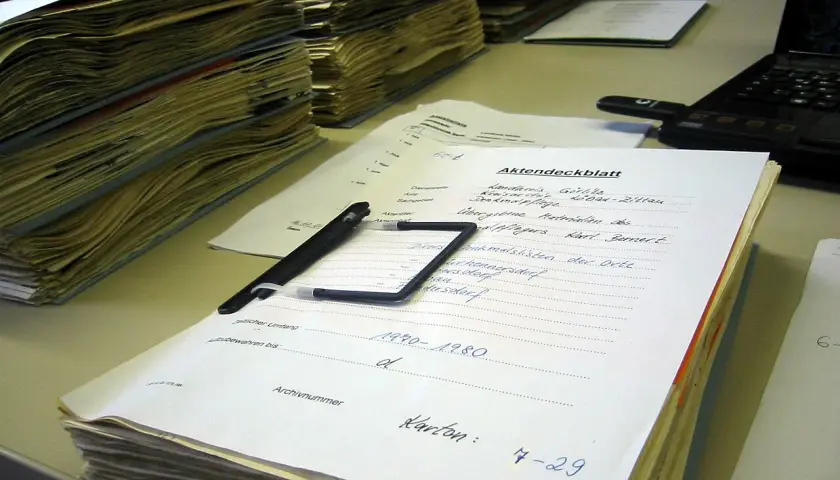 Stosy dokumentów, długopis i laptop na biurku. Na jednym z dokumentów widnieje napis "Akten-dossier co to", sugerujący porządkowanie akt.