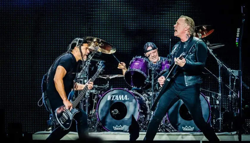 Duński zespół rockowy na scenie: basista z warkoczami, perkusista w fioletowym zestawie i gitarzysta w skórzanej kurtce.