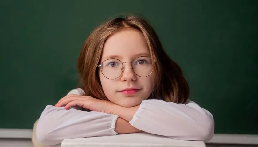 Dziewczynka w okularach opiera głowę na stosie książek przed zieloną tablicą.