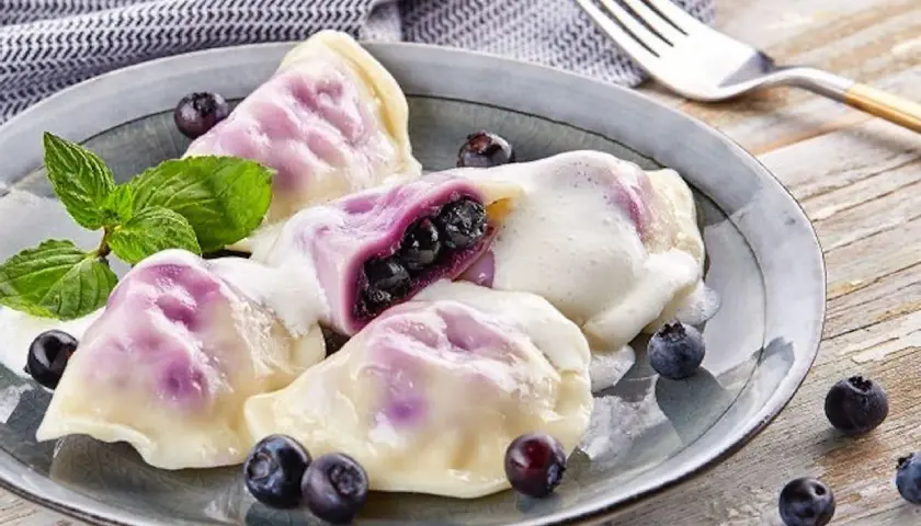 Pyszne pierogi z jagodami, polane śmietaną, ozdobione miętą i świeżymi owocami. Idealne na deser!