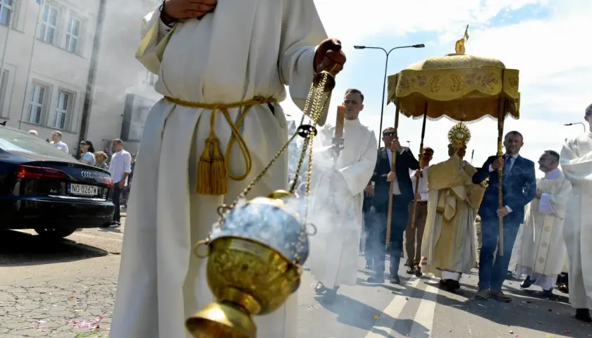 Procesja Bożego Ciała z kadzidłem i monstrancją. Uczestnicy w białych szatach idą ulicą.