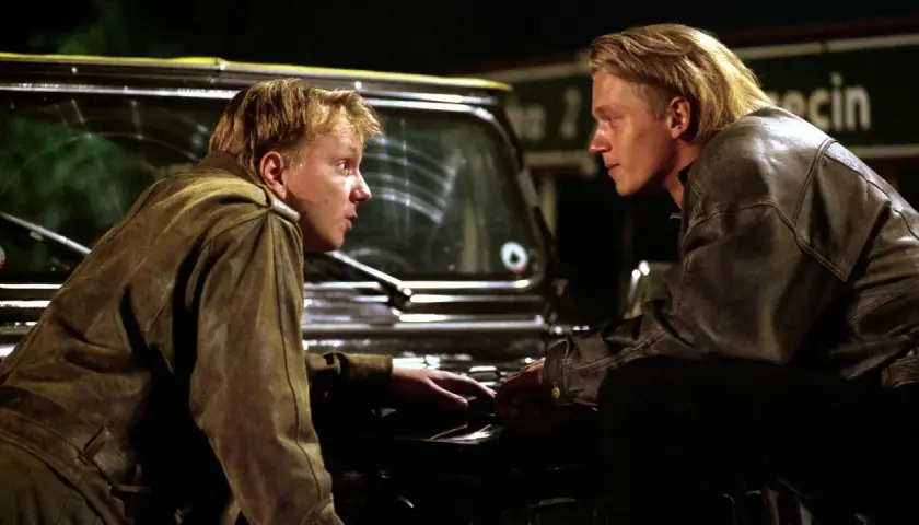 Dwóch młodych mężczyzn w skórzanych kurtkach, jak z filmów o mafii, pochyla się nad maską samochodu, prowadząc intensywną rozmowę.