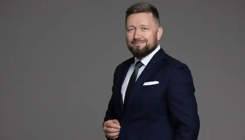 Portret mężczyzny w granatowym garniturze, białej koszuli i zielonym krawacie, z brodą i niebieskimi oczami.