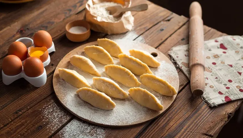 Surowe pierogi ułożone na desce, obok jajka, mąka i wałek.